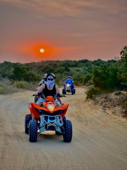 Two people riding ATVs on a dirt road with a sunset in the background. 1