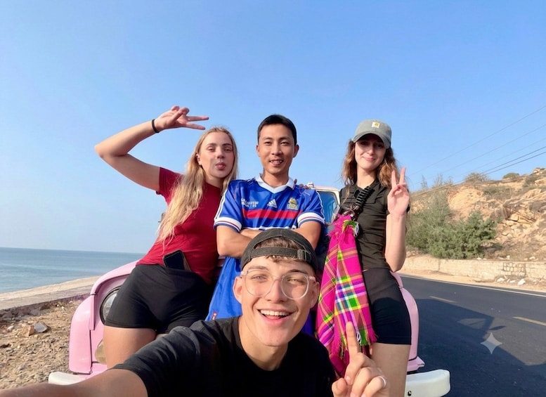 Four people on a pink scooter by the roadside with a clear sky and rocky terrain in the background. 1
