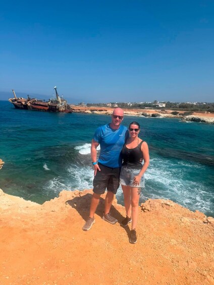A man and woman standing on a rocky shore with a shipwreck in the background. 5