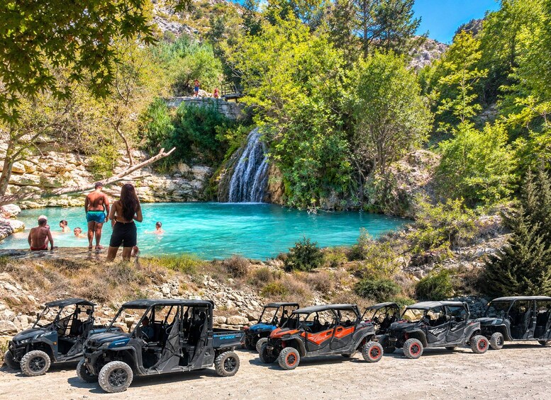 A turquoise pool surrounded by trees, a waterfall, and off-road vehicles parked in the foreground. 1