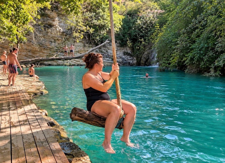 A person sitting on a wooden log in a turquoise pool, surrounded by greenery and rocky cliffs. 4