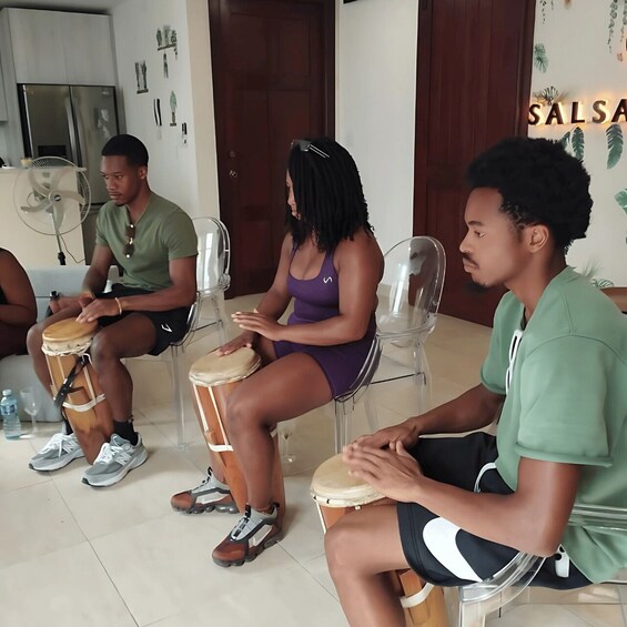 Four people seated on chairs, playing drums in a room with a 'Salsa' sign on the wall. 2