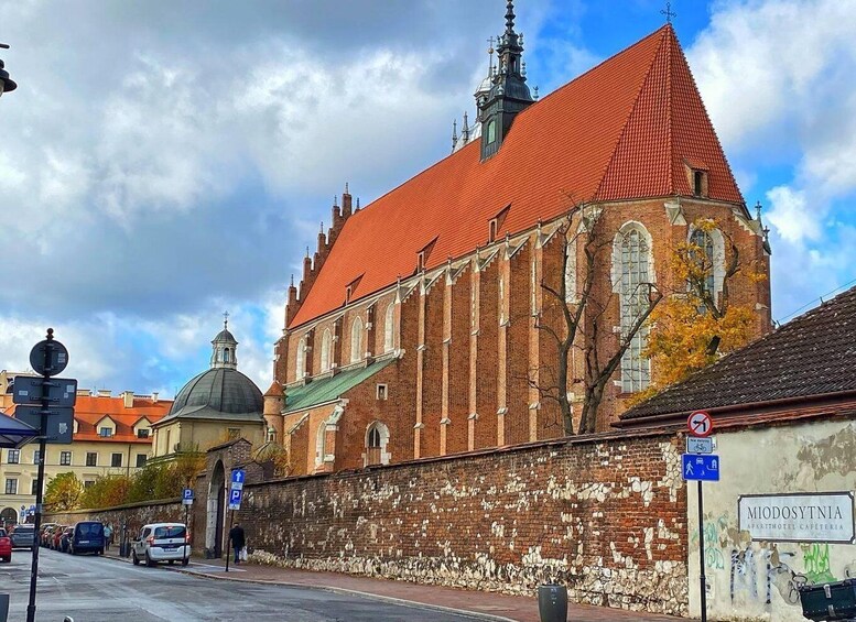A historic building with a red roof and a large dome, adjacent to a street with parked cars and a sign for 'Miodosytnia'. 3