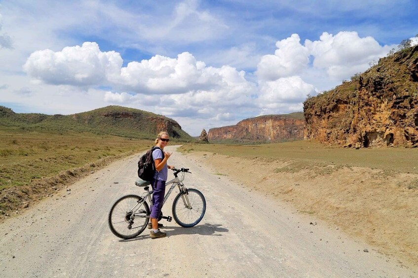 A person standing next to a bicycle on a dirt road with a mountainous landscape in the background. 1