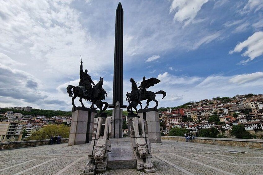 Een monument met twee standbeelden van bereden figuren aan weerszijden van een hoge obelisk, voor een achtergrond van een stadsgezicht met gebouwen en een bewolkte lucht. 4