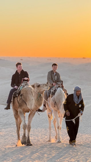 Three people riding camels in a desert at sunset. 1