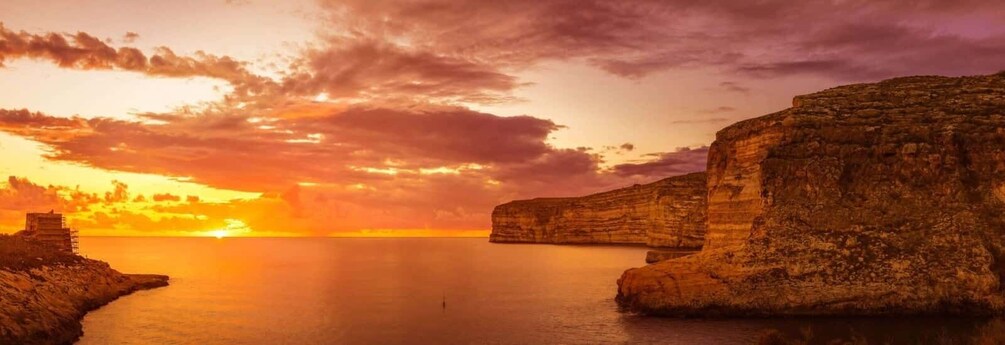 A sunset over a calm sea with cliffs and a small structure on the left. 5