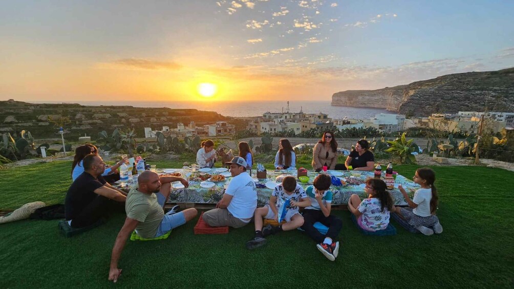 A group of people having a picnic on a grassy hill overlooking a coastal landscape during sunset. 3