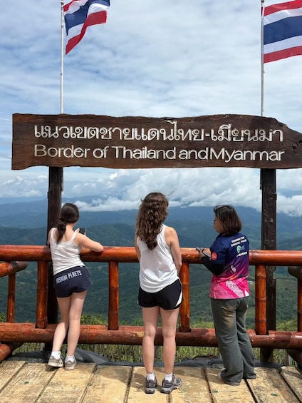 A wooden sign with text in English and another language, flags of Thailand and Myanmar, and three people standing on a wooden platform overlooking a mountainous landscape. 4