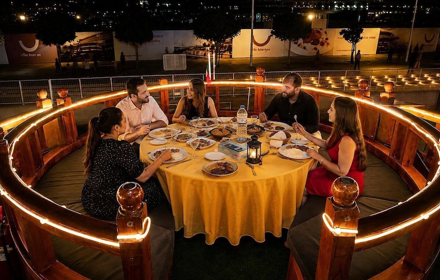 A group of people dining around a circular table with a yellow tablecloth, surrounded by wooden benches and illuminated by string lights. 5