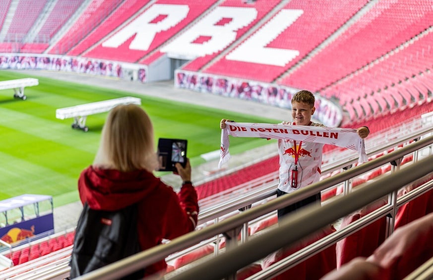 A person holding a scarf with text, standing in a stadium with red seats and a visible field. 5
