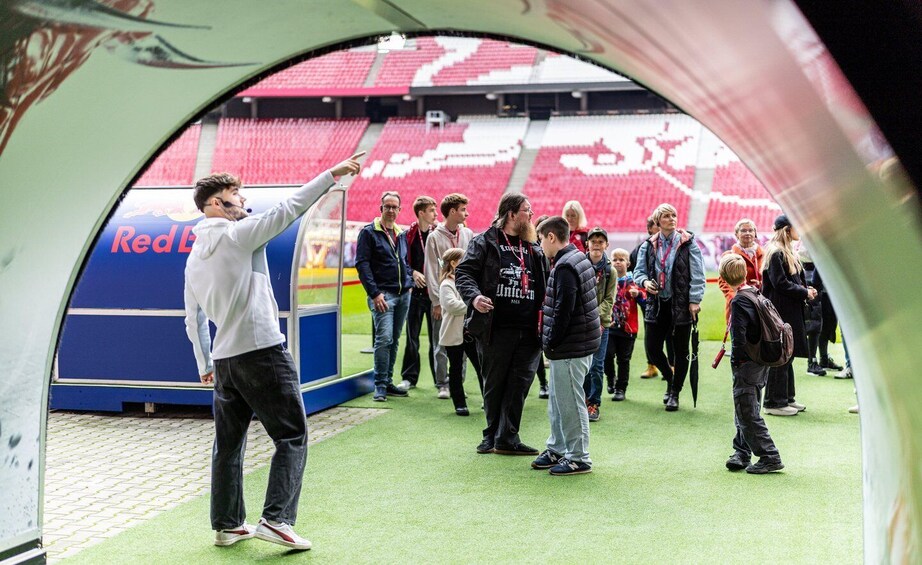 A group of people standing in a stadium with red seats and a Red Bull advertisement. 2