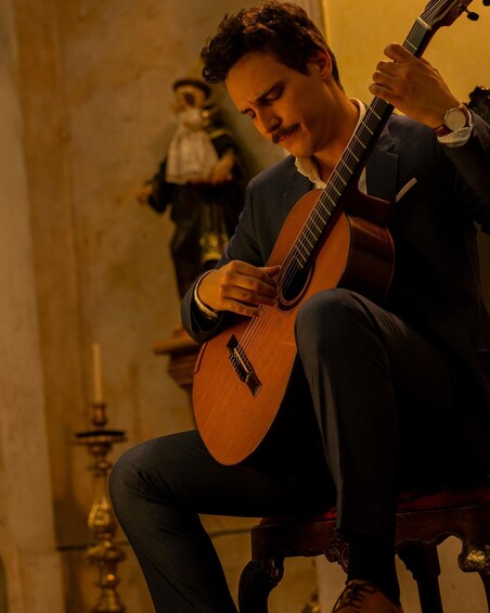 A person playing a guitar in a dimly lit room with a statue in the background. 3