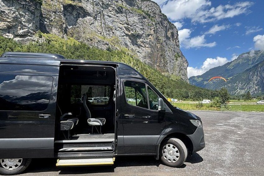 Ein schwarzer Van mit geöffneter Tür steht geparkt, mit Stufen, die zum Fahrzeug führen, und eine bergige Landschaft im Hintergrund. 3