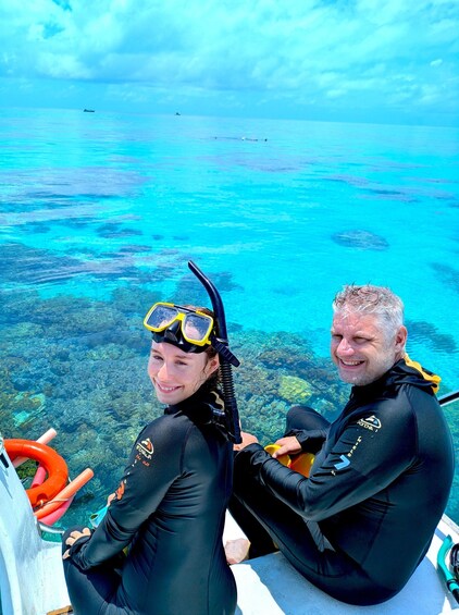 Two people in black wetsuits sitting on a boat with a clear view of the turquoise ocean and underwater landscape. 3