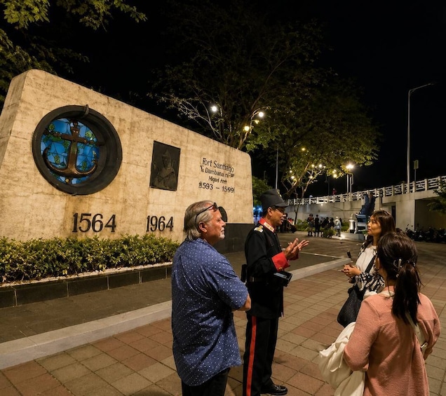 Escena nocturna con un monumento que presenta un vitral e inscripciones, rodeado de un área pavimentada con gente y un oficial uniformado. 5