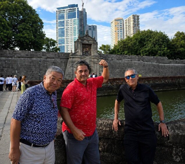 Three men posing by a waterfront with a historic building and modern skyscrapers in the background. 4