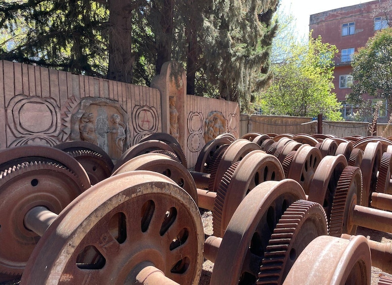 Large, rusted gears piled outdoors, with a decorative wall and trees in the background. 2