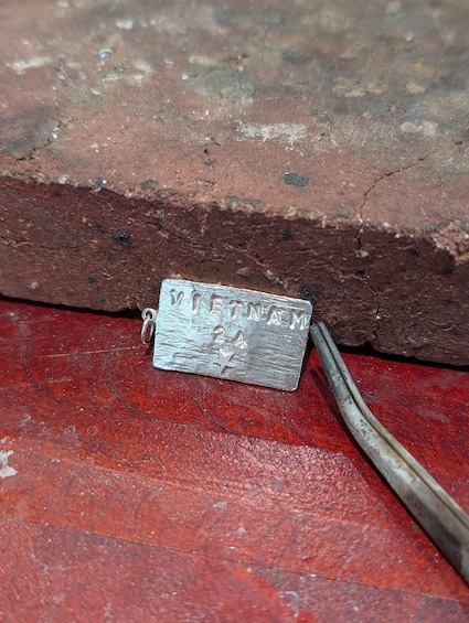 Metallinen tunnuslappu, johon on kaiverrettu teksti ”VIETNAM 24”, lepää punaisella pinnalla tiiliseinän halkeaman vieressä. 4