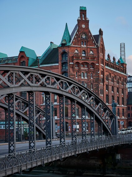 A steel bridge with ornate metalwork spanning a river. In the background, there is a large red brick building with a green roof and a clock tower. 5