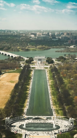 An aerial view of a large open plaza with a central monument, surrounded by a circular walkway and a reflecting pool. 2