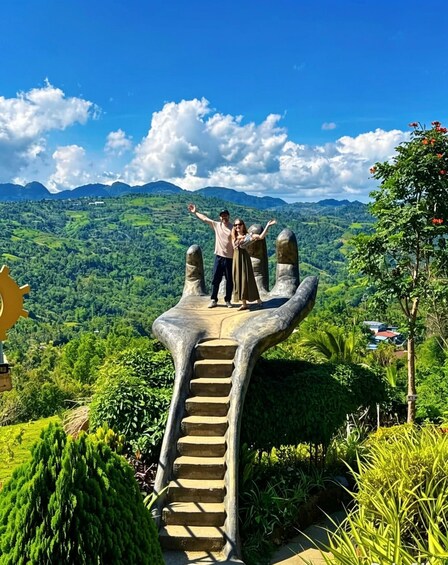 A couple standing on a hand-shaped staircase with a mountainous landscape in the background. 1