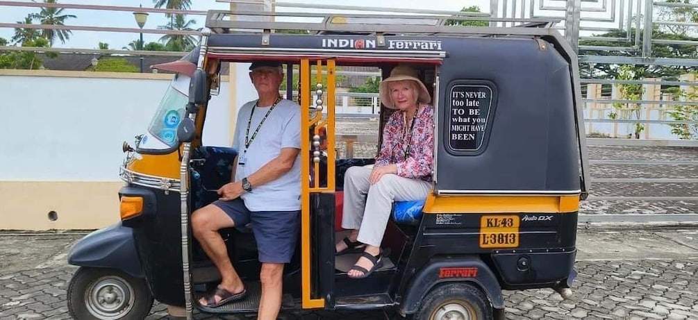 A man and a woman sitting in a yellow auto-rickshaw with a sign that reads "It's never too late to be what you might have been." 1