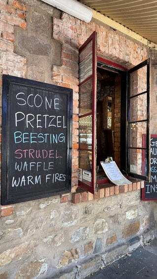 A brick building with a chalkboard sign listing items like scones, pretzels, and waffles. 1