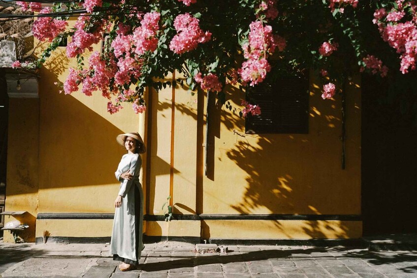 A person standing in front of a yellow building with pink flowers hanging from the wall. 2