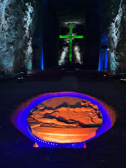 A cave interior with a green cross, benches, and a central illuminated sculpture. 1