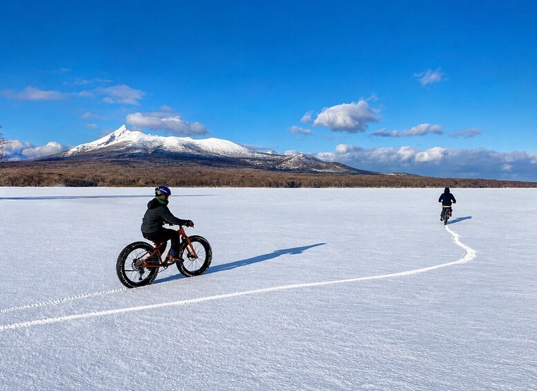 Two cyclists on a snowy flat surface with a mountain in the background. 5