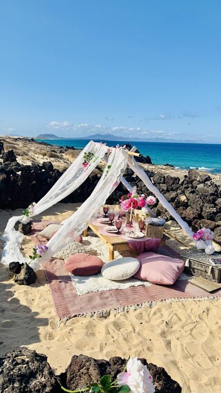 A beach picnic setup with a canopy, blankets, cushions, and a table with drinks and flowers. 5