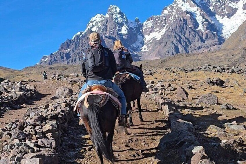 Due persone cavalcano cavalli su un sentiero roccioso con montagne innevate sullo sfondo. 2