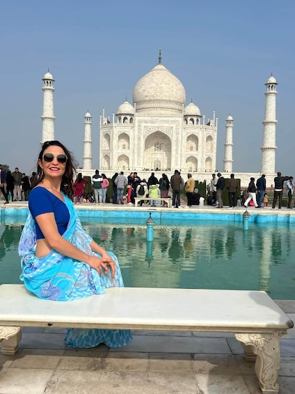 A person sitting on a bench in front of the Taj Mahal, with a pool and visitors in the background. 1