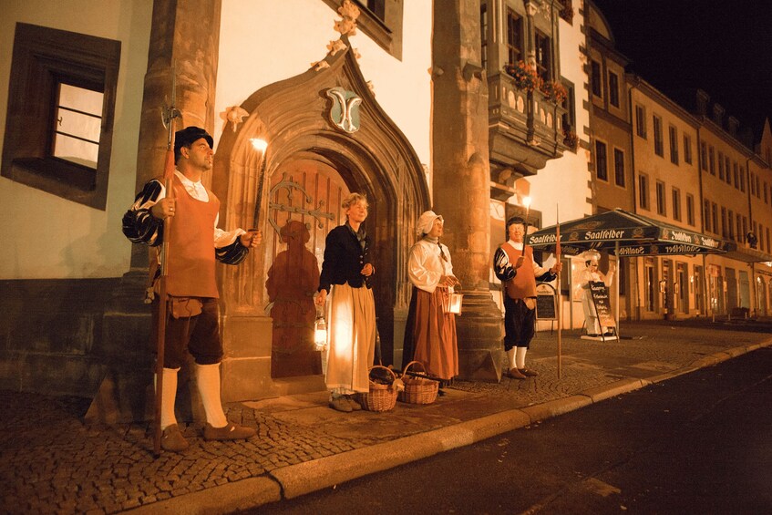 A historical reenactment with actors in period costumes outside a building with an ornate archway. 2
