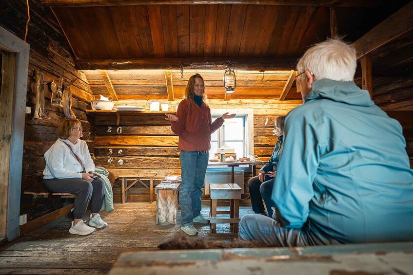 A rustic wooden cabin interior with a person standing and gesturing, three others seated on benches, and a window letting in natural light. 2