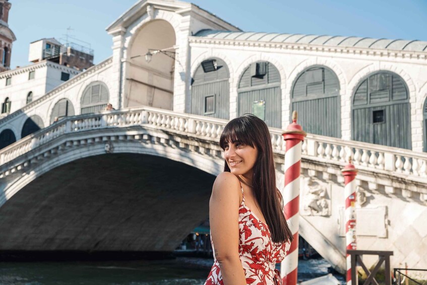 A woman in a floral dress standing in front of a historic bridge with arched windows and a lighthouse. 1