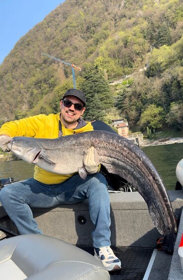 A person holding a large fish on a boat with a mountainous landscape in the background. 5