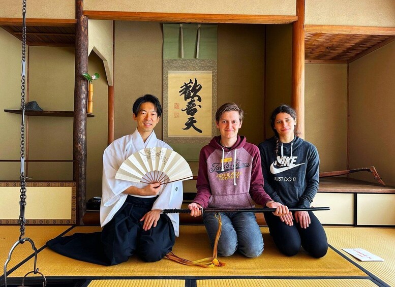 Three people sitting on tatami mats in a traditional Japanese room with a shoji screen and a hanging fan. 1
