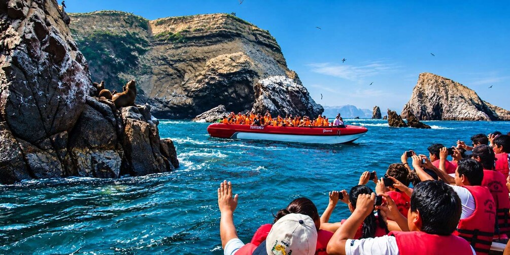 A group of people in red life jackets on a boat, with rocky cliffs and clear blue water in the background. 2