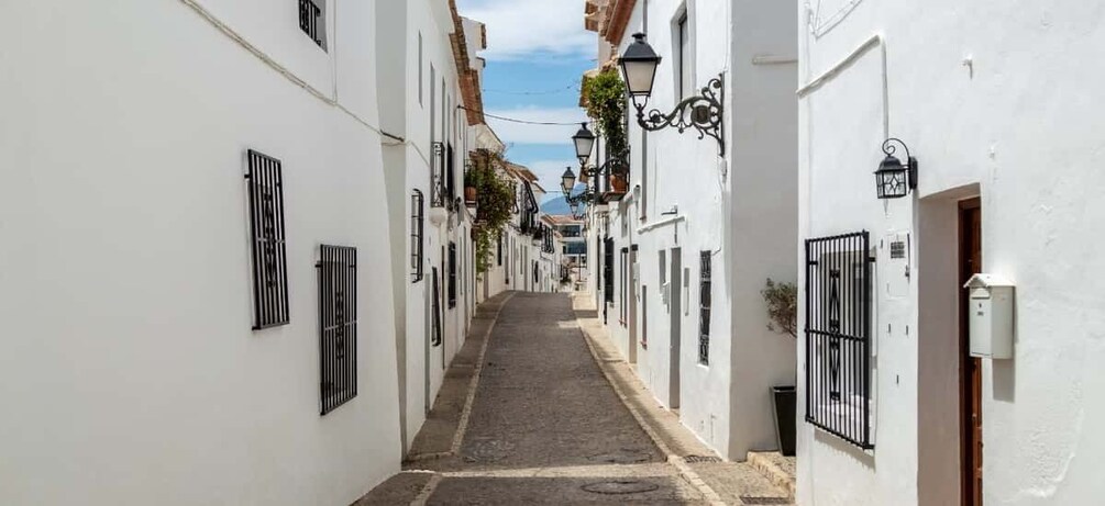 A narrow cobblestone street flanked by white buildings with black window bars and hanging plants. 3