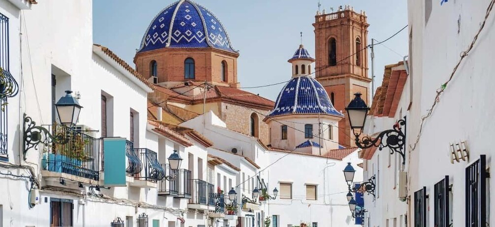 A narrow street with white buildings, blue domed churches, and a red bell tower. 1