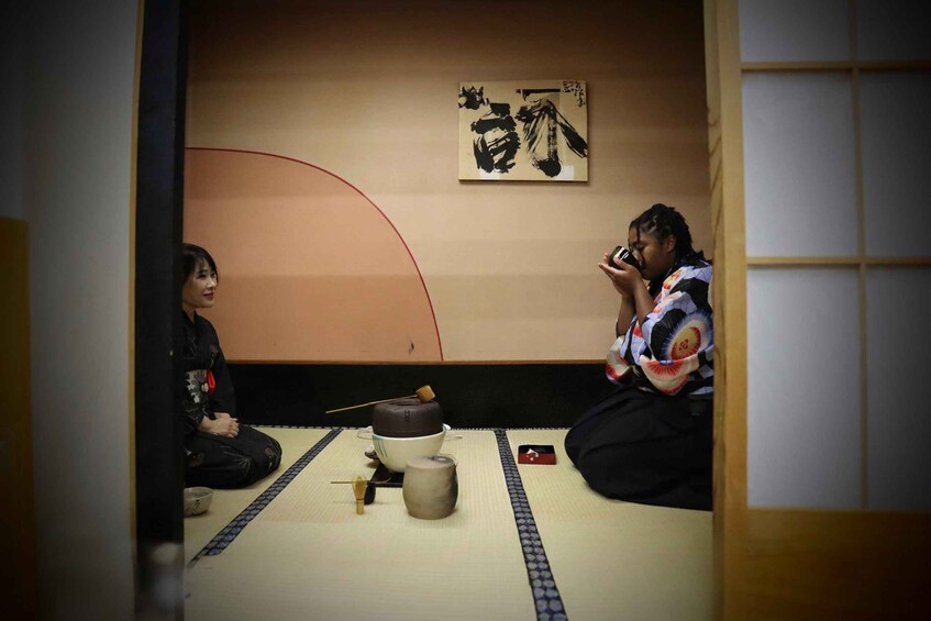 Two people sitting on tatami mats with a tea ceremony setup including a tea bowl, whisk, and sugar bowl. 5