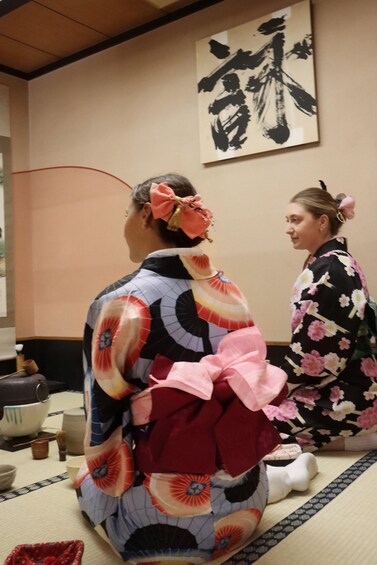Two individuals in traditional Japanese attire sitting on tatami mats with a tea ceremony setup in the background. 2