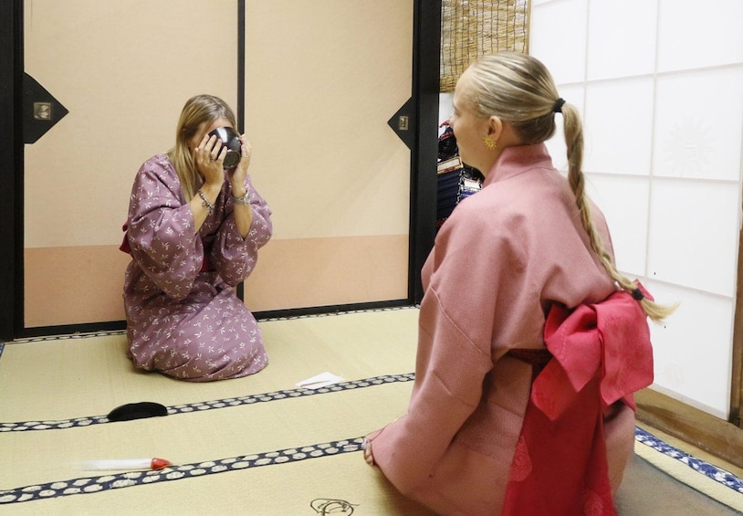 A person in a purple kimono taking a photo with a camera, another person in a pink kimono with a red sash, and a traditional Japanese tatami mat floor. 2