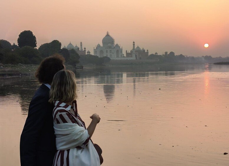 A couple standing by a river with the Taj Mahal in the background during sunset. 1