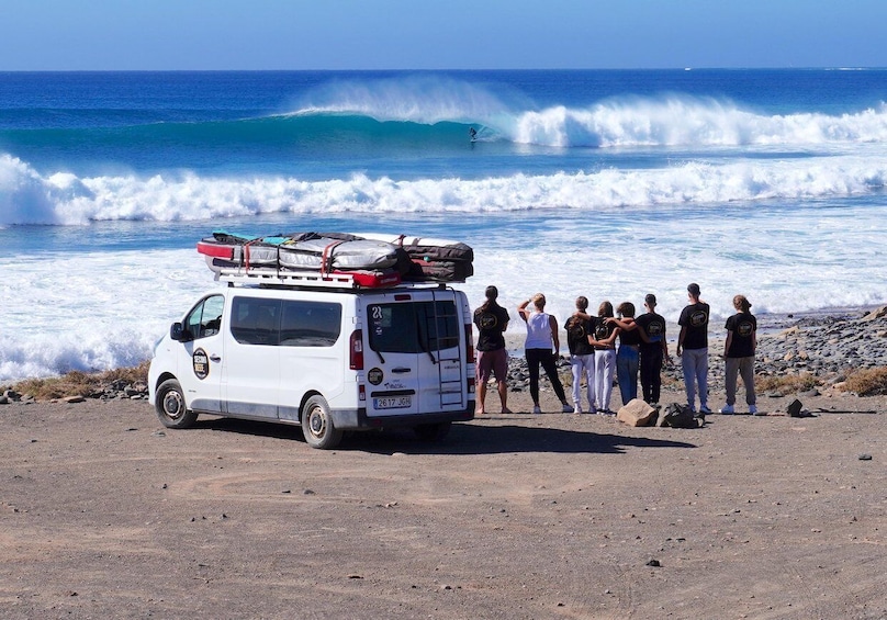 A white van parked on a beach with people standing nearby, a surfer riding a wave in the ocean, and a clear blue sky. 5
