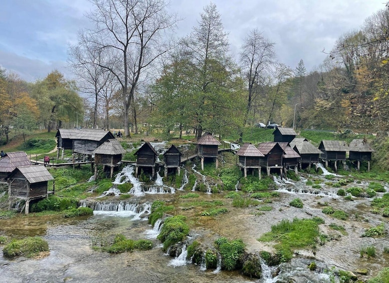 Wooden structures on stilts over a flowing stream, surrounded by trees and greenery. 1