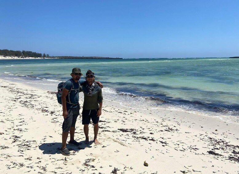 Two people standing on a sandy beach with clear blue water and a distant shoreline. 4