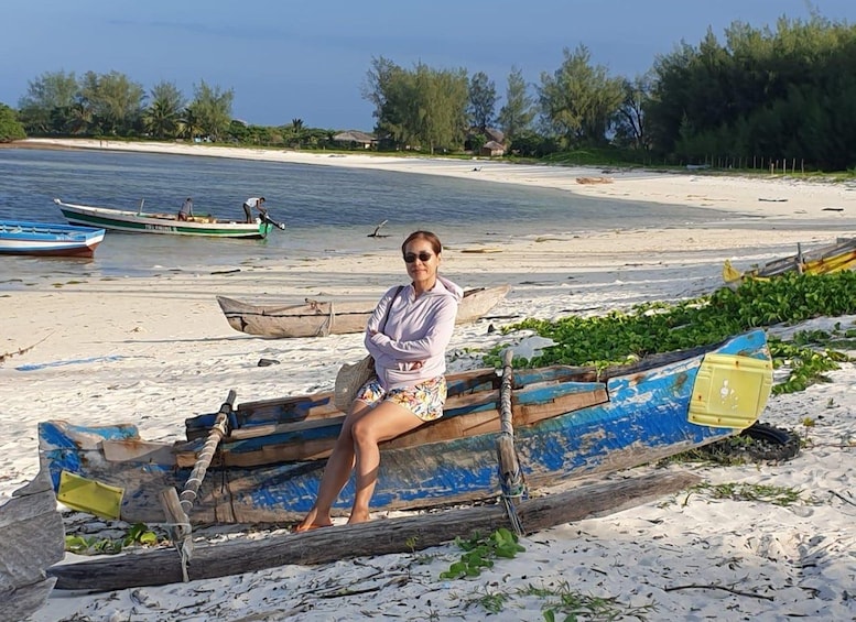 A person sitting on a wooden boat on a sandy beach with other boats and vegetation in the background. 2